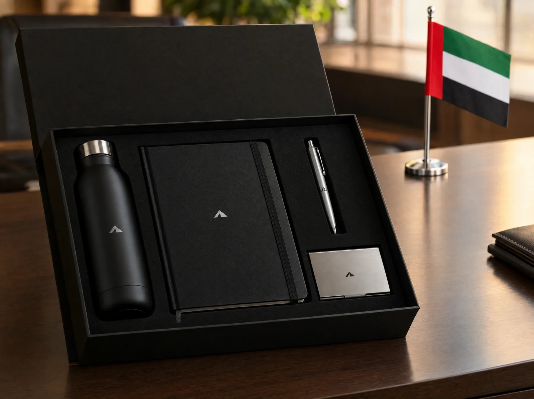 Black gift box with a steel bottle, notebook, pen and card holder, with a UAE desk flag on the desk behind